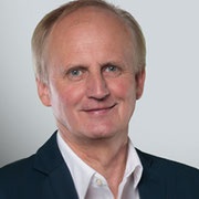 Ein Mann mit kurzen blonden Haaren, der ein weißes Hemd und einen dunklen Anzug trägt, lächelt in die Kamera. Er hat einen fröhlichen Ausdruck.