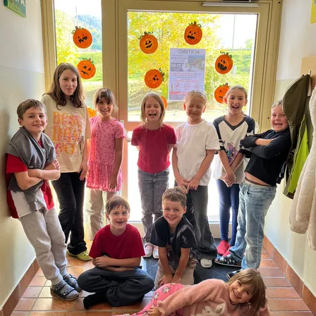 Eine Gruppe von Kindern posiert für ein Foto in einem Flur mit einer Tür im Hintergrund. Die Tür hat eine Halloween-Dekoration mit Kürbissen und einem Schild.