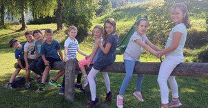 Eine Gruppe von Kindern sitzt auf einem Holzstamm im Freien. Sie lächeln und posieren für ein Foto. Im Hintergrund befinden sich grüner Rasen und Bäume.