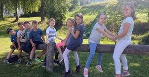 Eine Gruppe von Kindern sitzt auf einem Holzstamm im Freien. Sie lächeln und posieren für ein Foto. Im Hintergrund befinden sich grüner Rasen und Bäume.