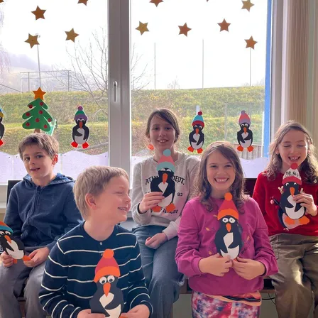 Eine Gruppe von Kindern, die in einem Klassenzimmer sitzen und Pinguin-Ausschnitte halten, lächeln mit einem festlichen Hintergrund aus Weihnachtsbäumen und Sternen.