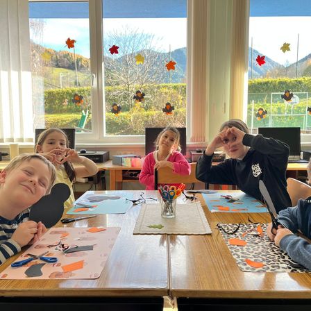 Fünf Kinder sitzen an einem Tisch und basteln. Papiere und Scheren sind um sie herum verstreut. Ein Mädchen in Pink hält Scheren und ein anderes Kind hält einen schwarzen Gegenstand. Hinter ihnen gibt es ein großes Fenster mit Blick auf Bäume und Berge.