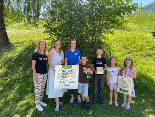 Gruppenfoto von Erziehern, Kindern und Eltern vor einem Baum mit einem Schild, das Natur-Garten Schule sagt.