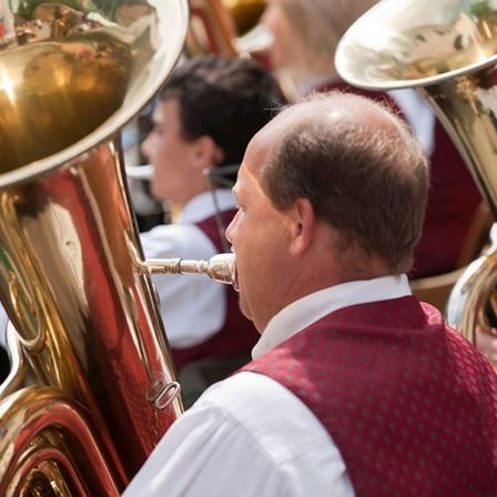 Ein Mann spielt ein Blasinstrument während einer musikalischen Aufführung. Er trägt eine rote Weste. Hinter ihm spielen andere ähnliche Instrumente.