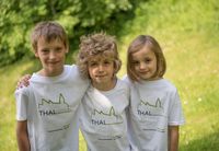 Drei Kinder stehen zusammen auf einem Rasenfeld und tragen passende weiße T-Shirts mit dem Wort 'THAL' in Grün auf der Vorderseite.
