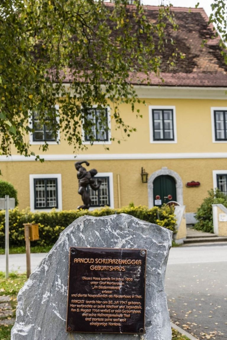 Ein gelbes Haus mit Fenstern und einer Statue davor. Eine Tafel ist auf einem Felsen vor dem Haus angebracht.