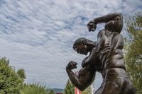 Eine Bronzestatue eines muskulösen Mannes, der seinen Arm anspannt, mit einem bewölkten Himmel und Bäumen im Hintergrund.