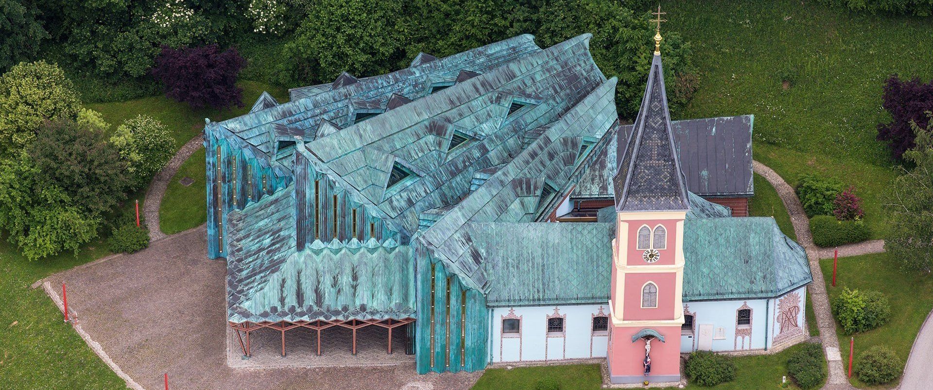 Ein Luftbild eines modernen Gebäudes mit einem grünen Dach und weißen Wänden. Es hat einen spitzen Turm mit einer Uhr und einem Kreuz oben.