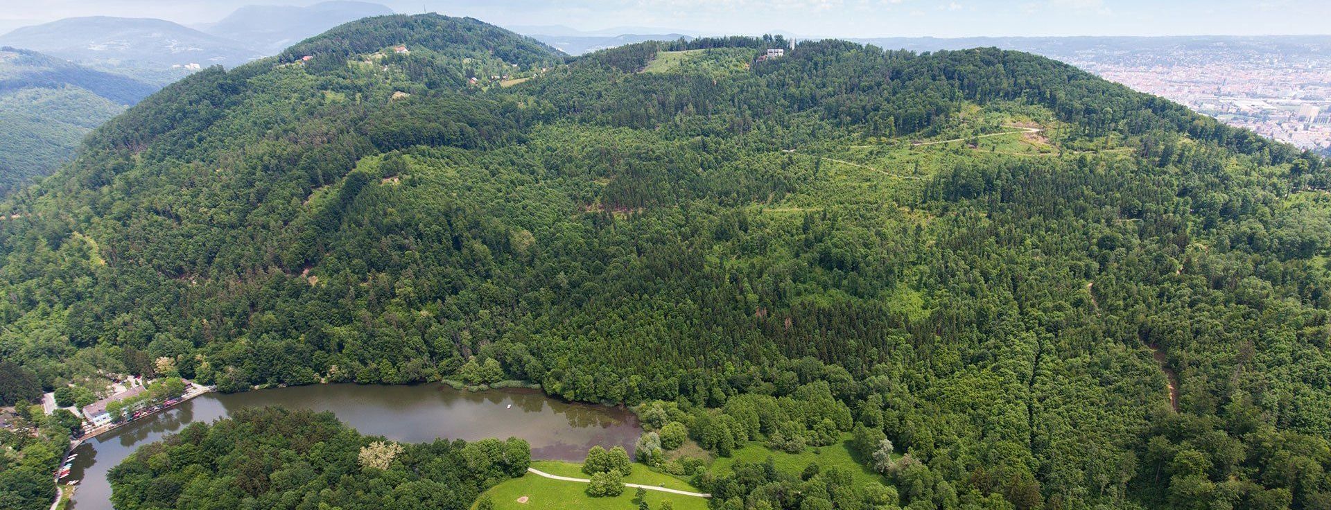 Luftaufnahme eines dichten Waldes mit einem Fluss, der sich schlängelt. Berge in der Ferne und ein Gebäude auf der Bergkuppe.