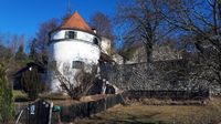 Ein altes weißes Gebäude mit braunem Dach und grünen Fensterläden steht vor einem klaren blauen Himmel. In der Nähe befinden sich eine Steinmauer und ein Holzzaun.