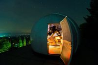 Ein Observatorium bei Nacht mit einer kuppelförmigen Struktur, offener Tür und Teleskop. Die Sterne sind sichtbar oben, und eine Stadtsilhouette ist in der Ferne zu sehen.