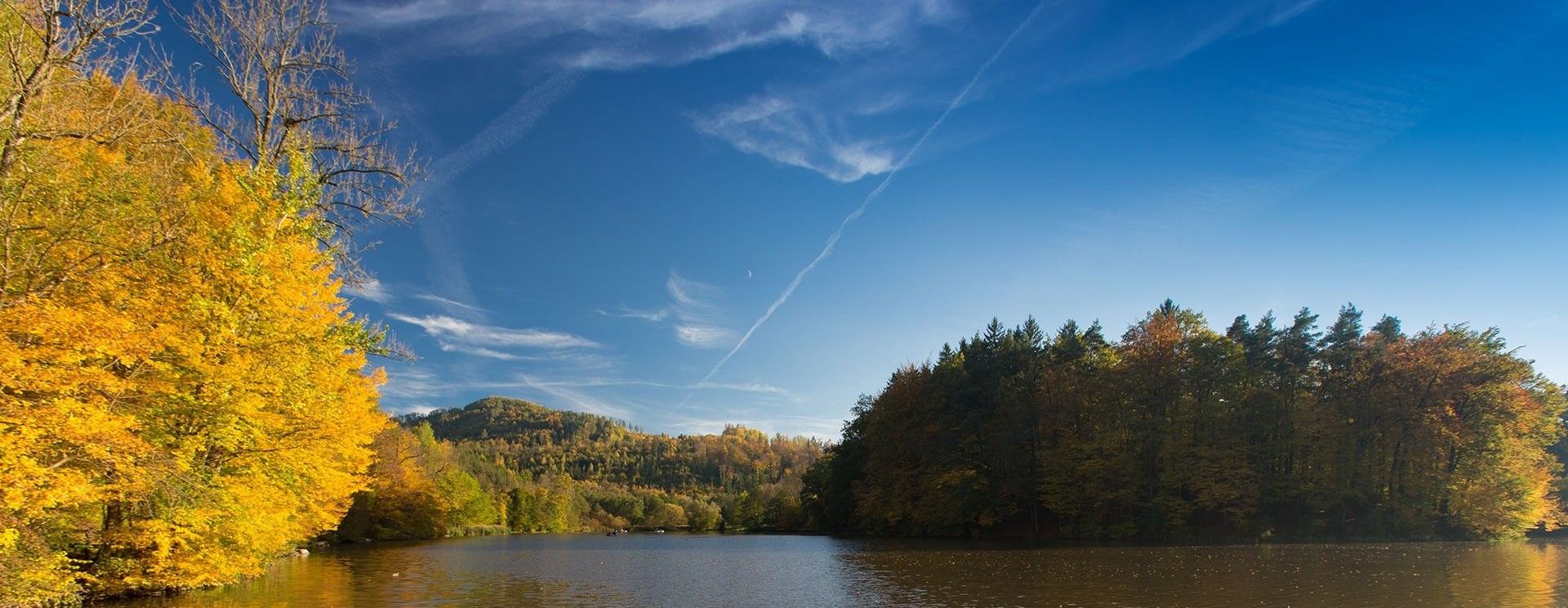 Ein ruhiger See mit blauem Himmel, einem Berg im Hintergrund und Bäumen mit verschiedenfarbigen Blättern. Ein Boot ist im Wasser.