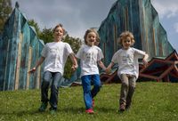 Drei Kinder in passenden weißen T-Shirts laufen fröhlich auf einem Grasfeld, mit einem einzigartigen grünen Gebäude im Hintergrund.