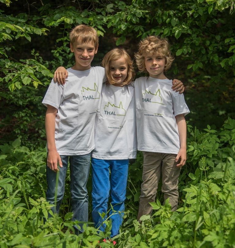 Drei Kinder stehen in einem üppigen grünen Feld und tragen passende weiße T-Shirts mit einem grünen Logo. Sie lächeln und posieren für ein Foto.
