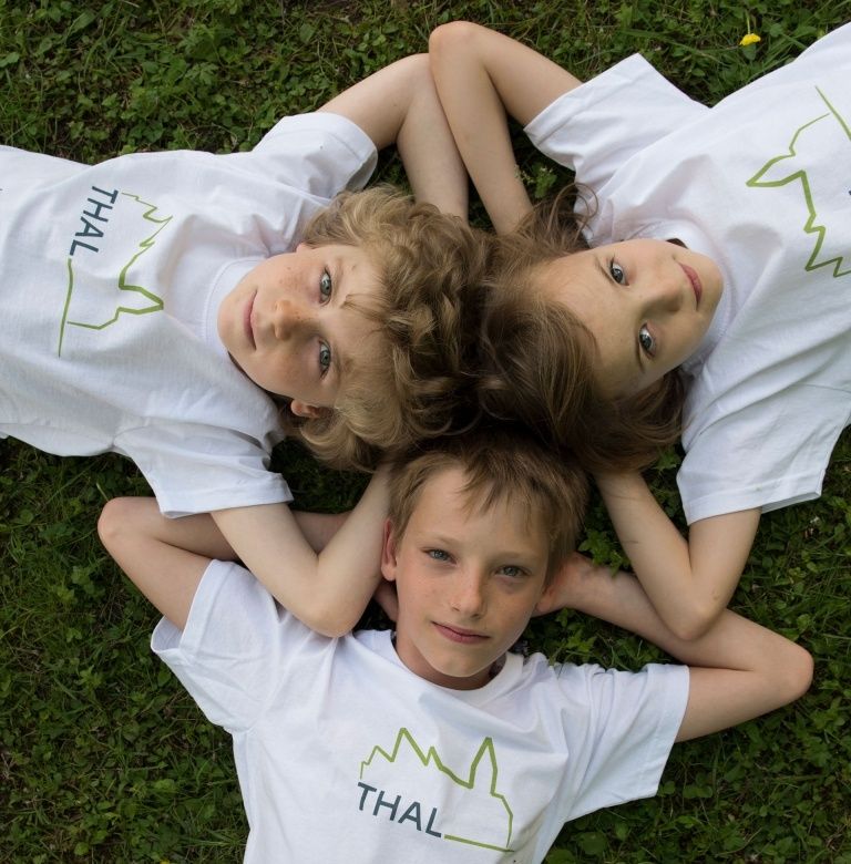 Drei Kinder liegen auf einem Grasfeld und tragen weiße T-Shirts mit dem Wort 'Thal' und einem grünen Bergdesign. Sie scheinen für ein Foto zu posieren.