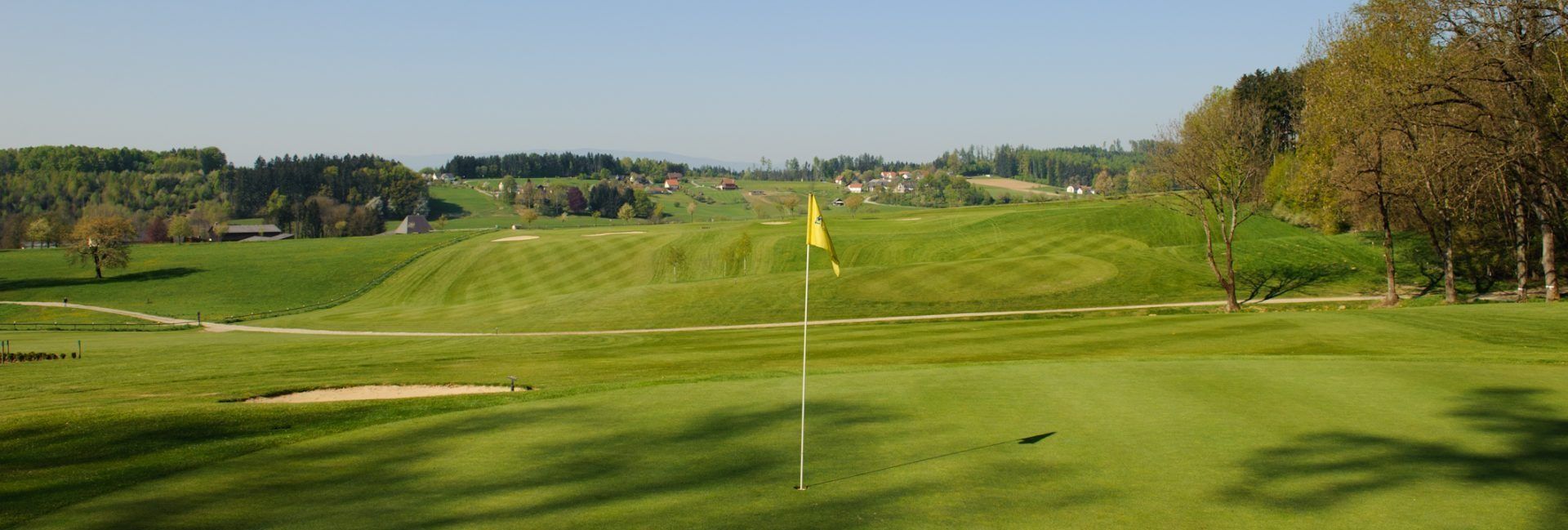 Ein Golfplatz unter einem klaren Himmel, mit einer gelben Fahne, die das Loch markiert. Das Feld ist üppig, und Häuser und Bäume sind in der Ferne.