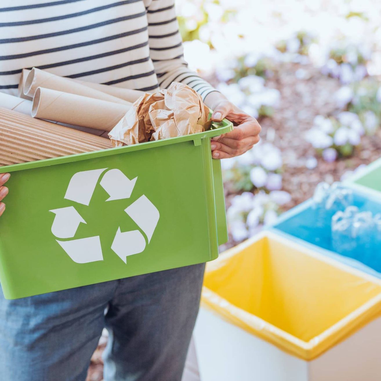 Eine Person in einem gestreiften Shirt hält einen grünen Recyclingbehälter mit recycelbaren Gegenständen. Dahinter befinden sich drei Behälter: gelb, blau und grün.