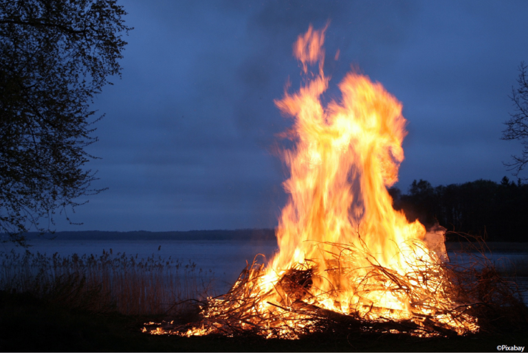 Ein Lagerfeuer brennt bei Nacht am See, umgeben von Ästen und trockenem Gras.