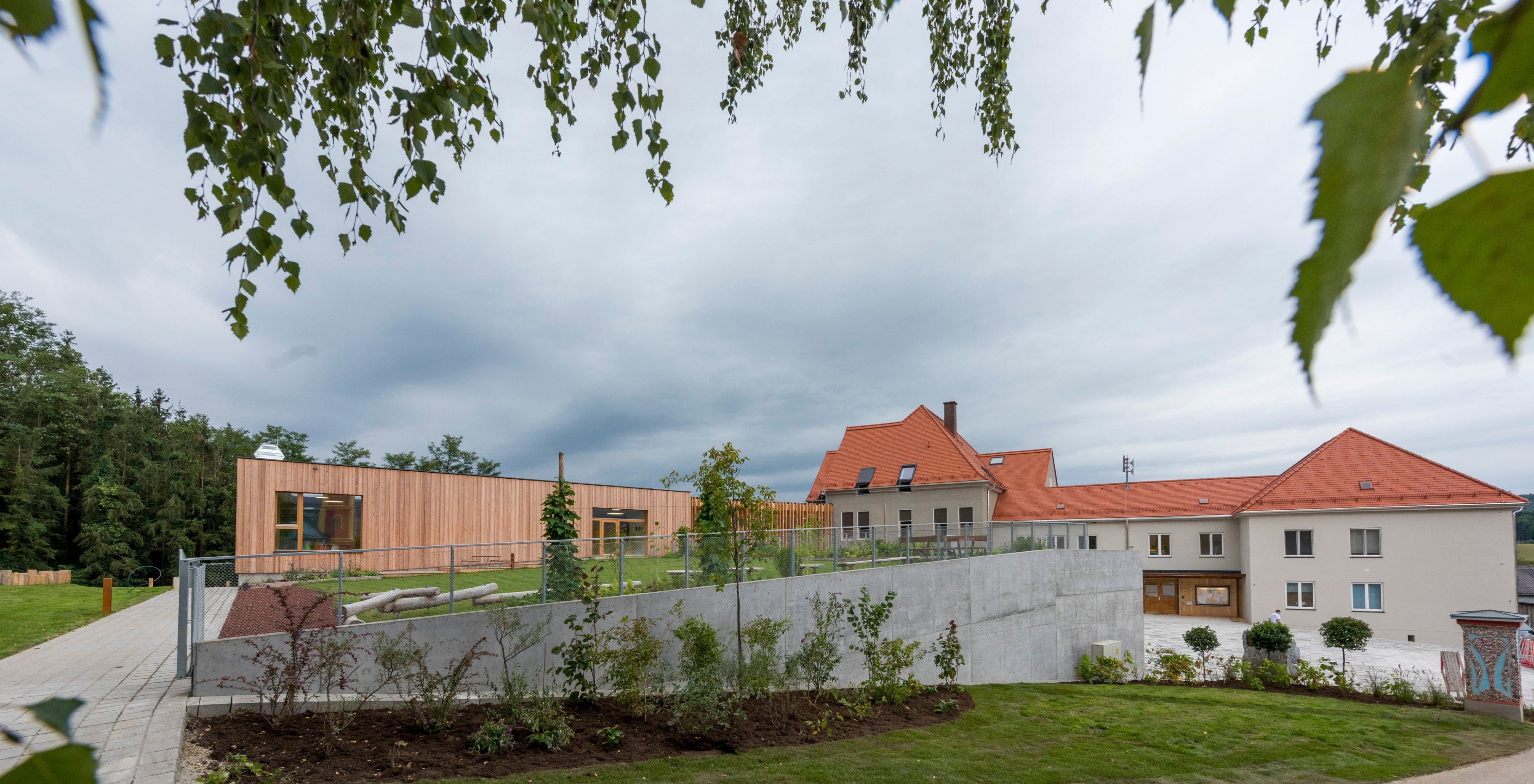 Ein modernes Gebäude mit einem roten Dach und einer Betonmauer davor. Das Gebäude ist von einem Rasenbereich mit Pflanzen und Bäumen umgeben.