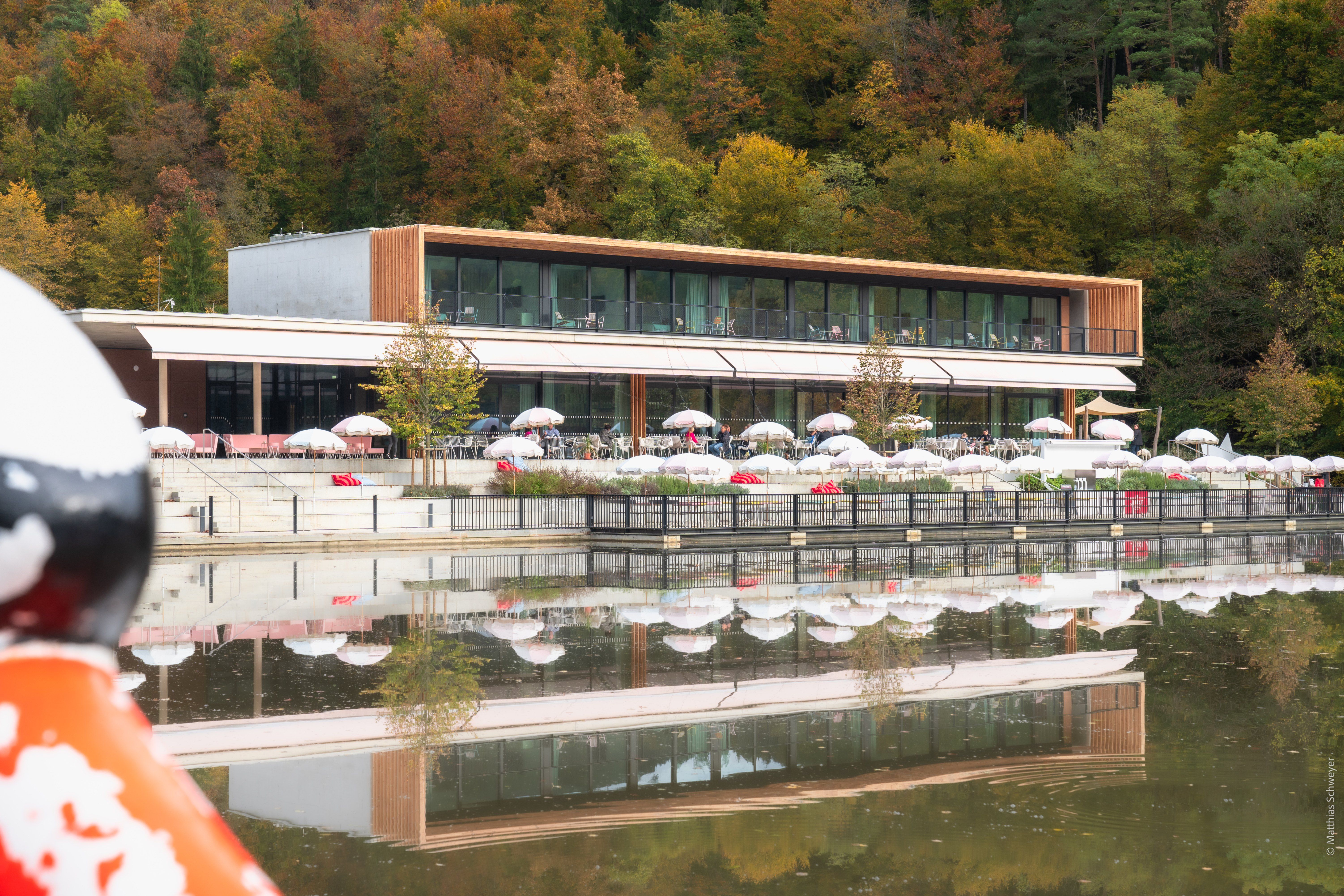 Ein modernes Gebäude mit Glaswänden und Holzakzenten spiegelt sich im Wasser. Viele weiße Sonnenschirme sind draußen aufgestellt.