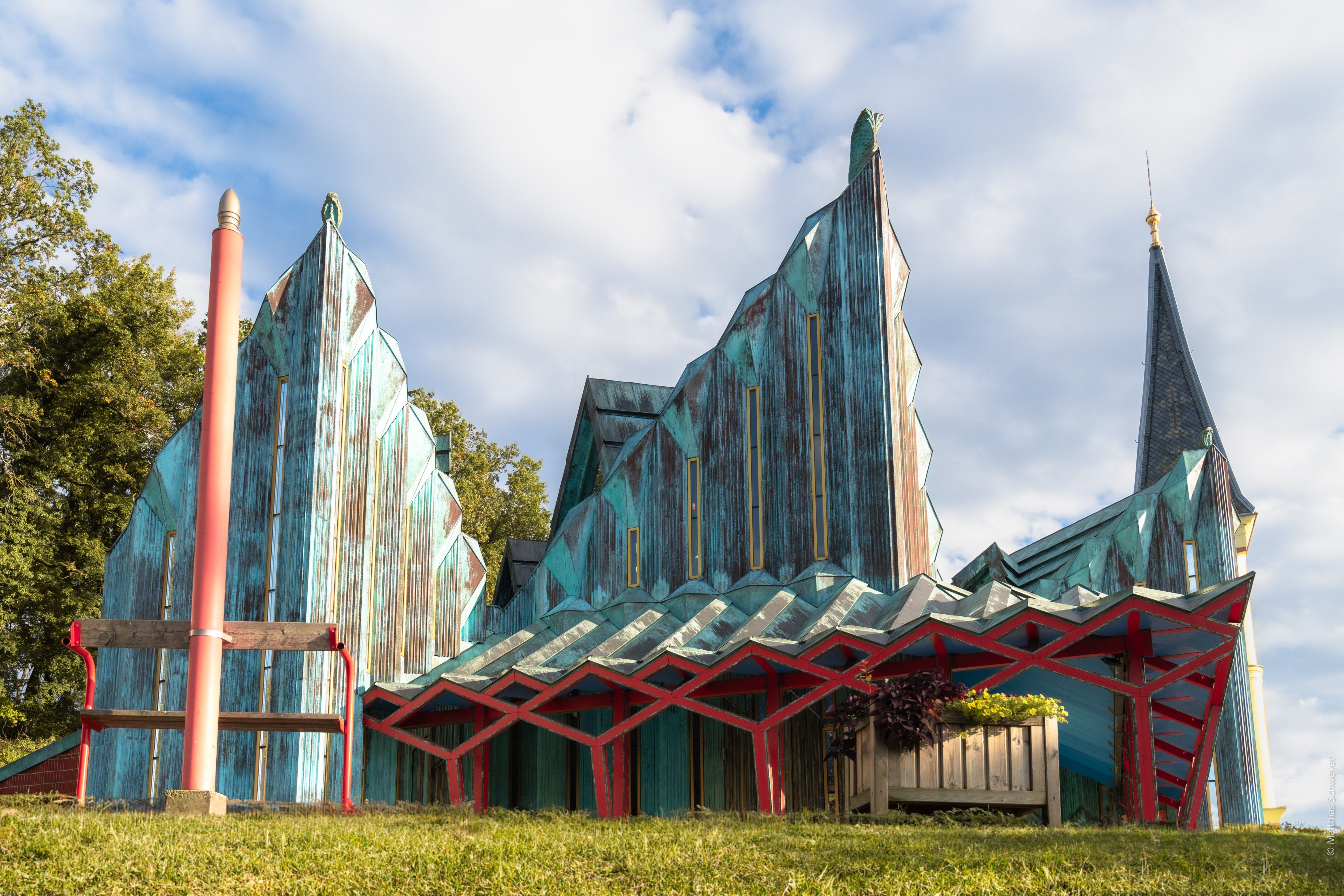 Ein einzigartiges, art-deco inspiriertes Gebäude mit türkisen und roten Akzenten steht auf einer Wiese unter einem bewölkten Himmel.