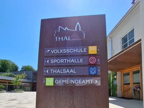 Ein braunes Schild mit weißen Texten und Symbolen weist auf Volksschule, Sporthalle, Thalsaal und Gemeindeamt hin. Jede Richtung hat ein entsprechendes Symbol.