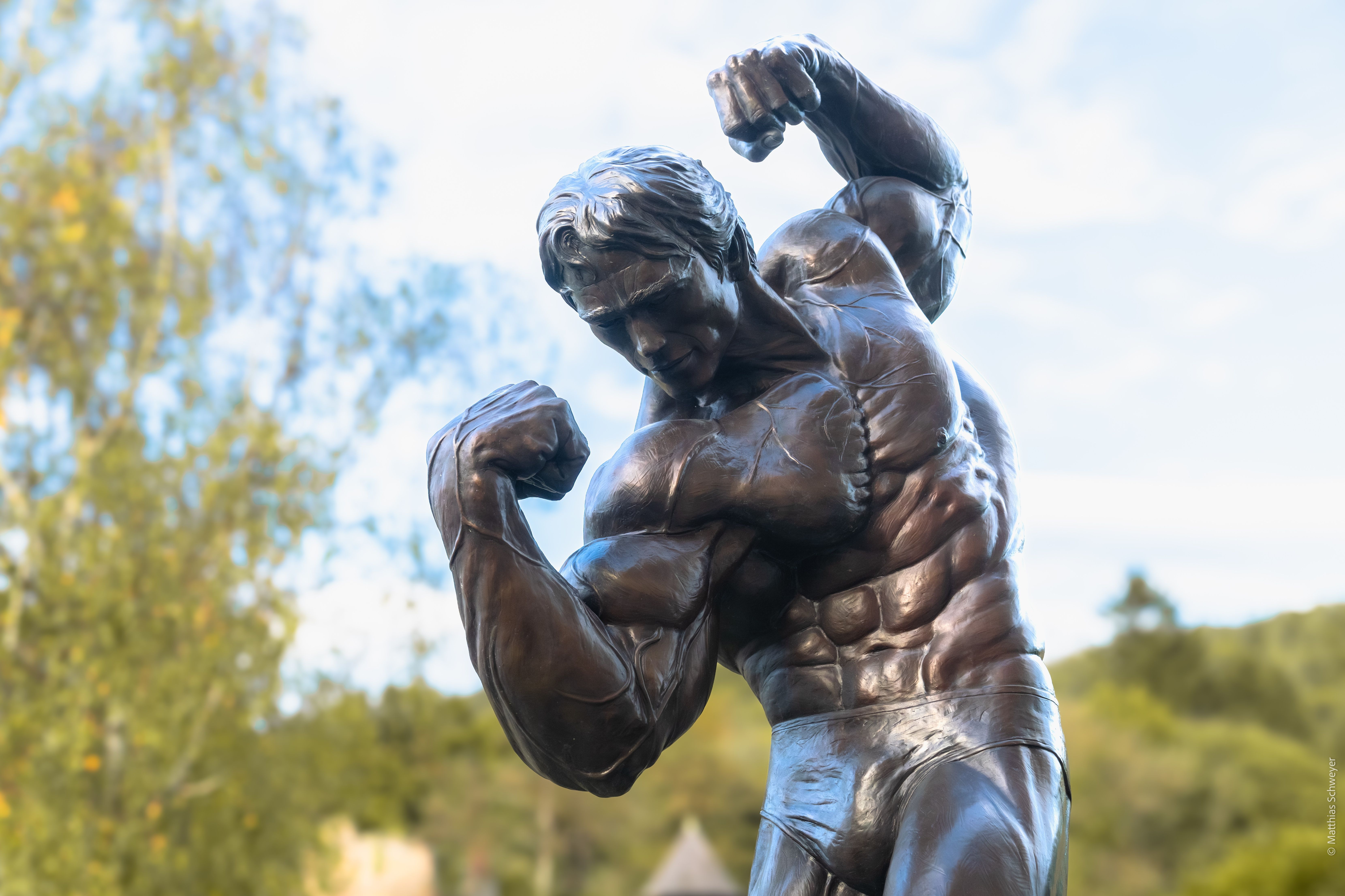 Eine Bronzestatue eines muskulösen Mannes, der seine Bizeps und Trizeps anspannt, steht im Freien mit klarem Himmel und Bäumen im Hintergrund.