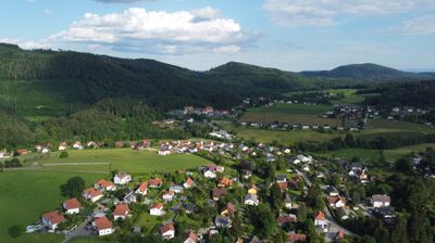 Luftaufnahme eines kleinen Dorfes zwischen grünen Hügeln und üppigen Feldern unter einem teilweise bewölkten Himmel.