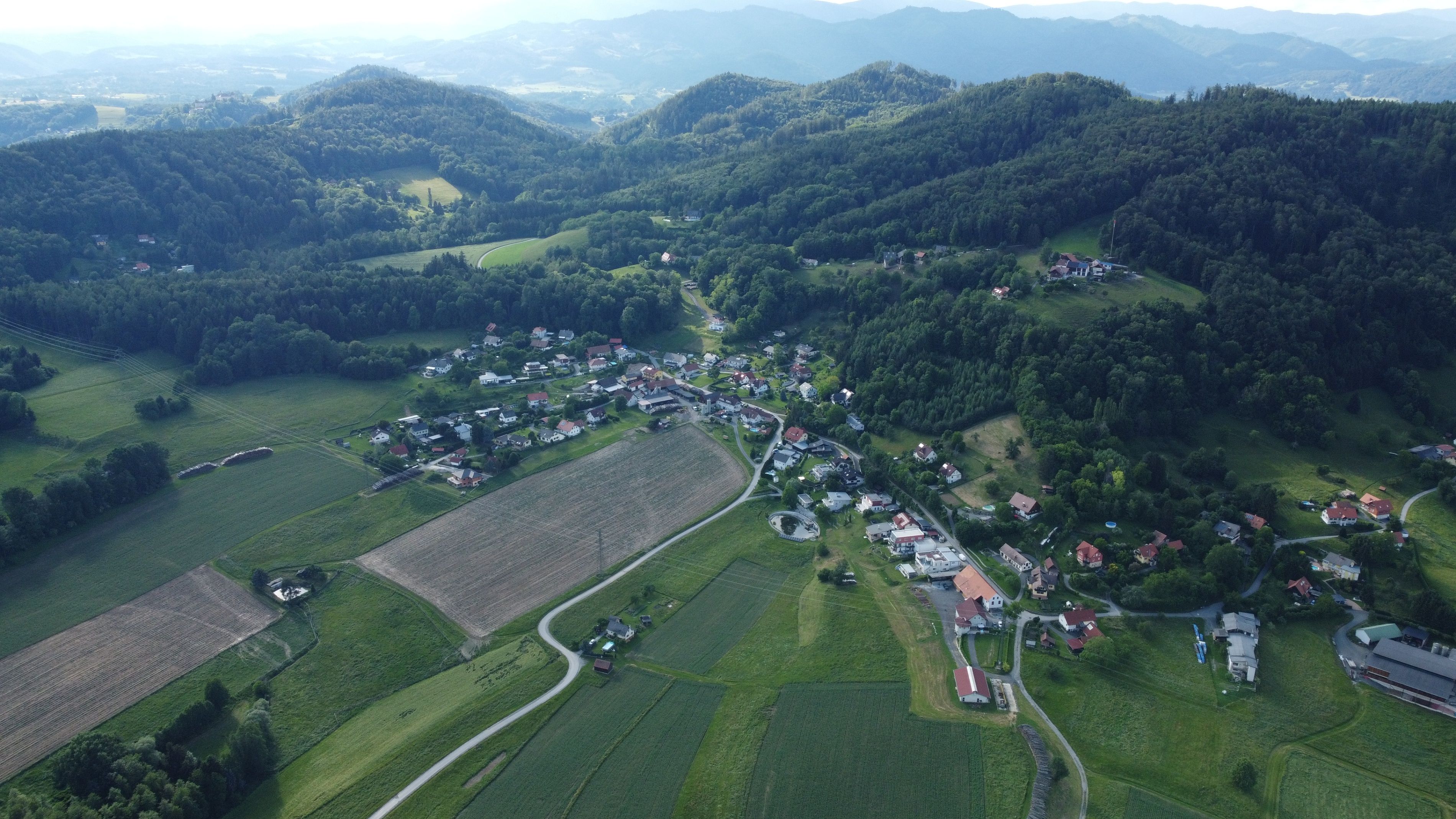 Luftaufnahme eines ländlichen Dorfes in einem Tal, umgeben von üppigen grünen Feldern und bewaldeten Hügeln.