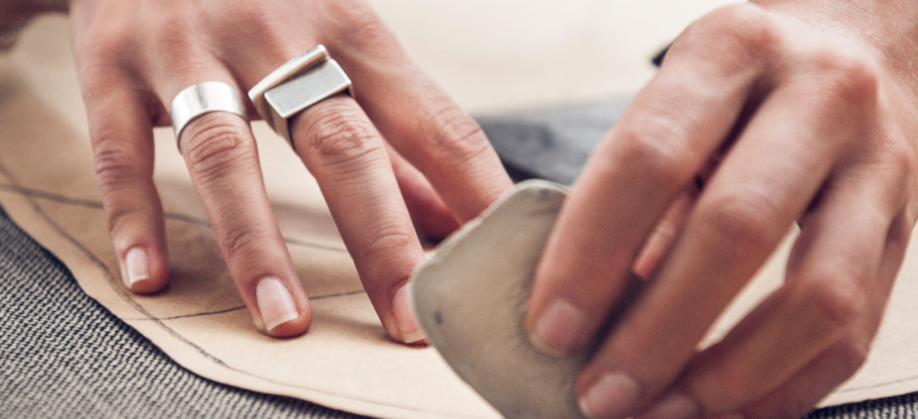 Eine Hand mit zwei silbernen Ringen hält ein Stück graues Material, möglicherweise Leder, und arbeitet daran.