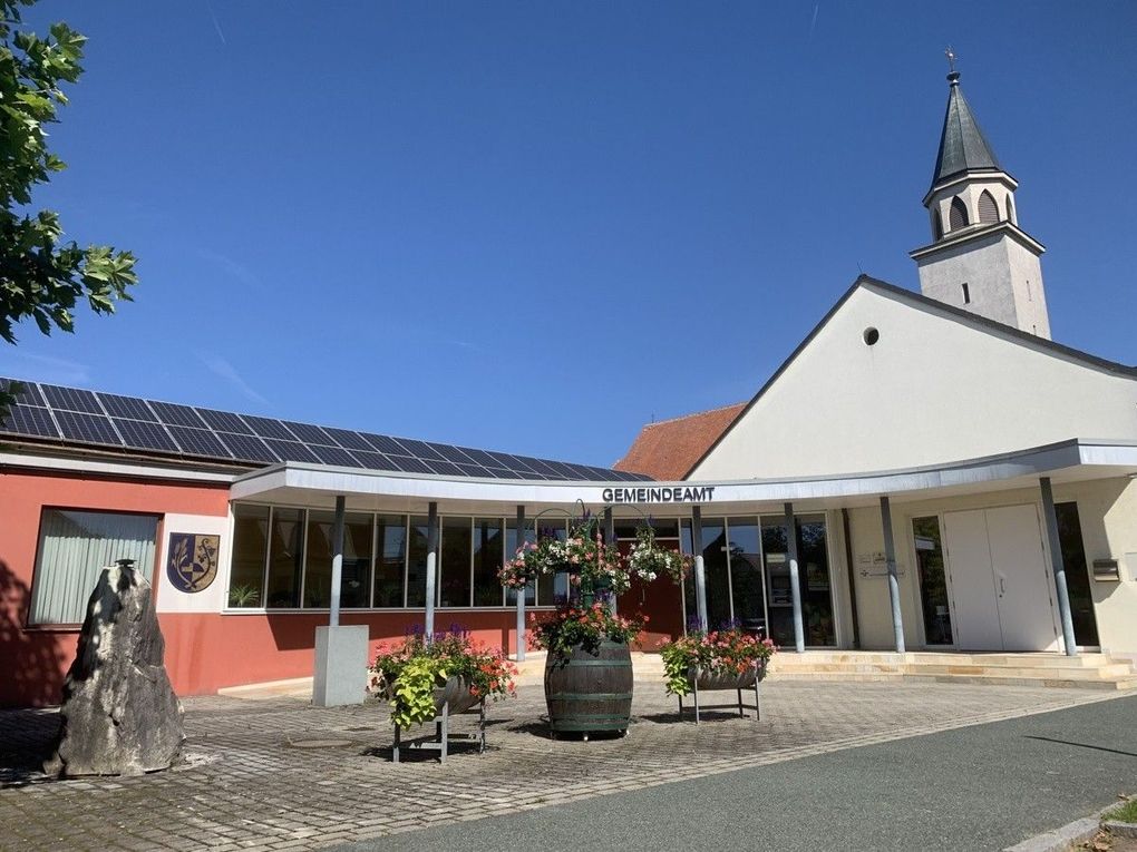  Abschnitt Bild: Ein Gebäude mit Solarpanelen auf dem Dach und einem Turm, davor mehrere Pflanzen und Blumen.