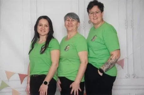 Drei Frauen in grünen T-Shirts stehen zusammen vor einem weißen Hintergrund mit bunten Wimpelgirlanden. Sie lächeln und scheinen für ein Foto zu posieren.