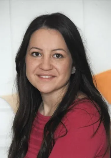 Eine lächelnde Frau mit langen schwarzen Haaren in einem roten Shirt posiert vor einem weißen und orangefarbenen Hintergrund.