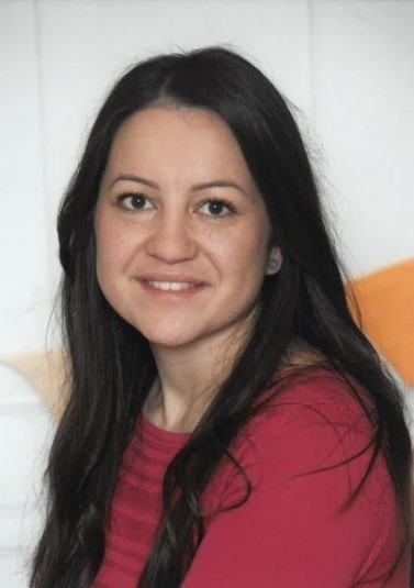 Eine lächelnde Frau mit langen schwarzen Haaren in einem roten Shirt posiert vor einem weißen und orangefarbenen Hintergrund.