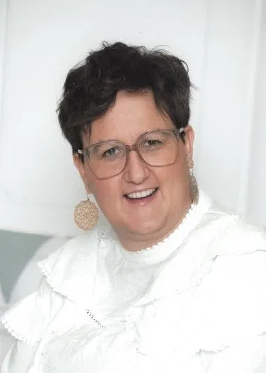 Eine Frau mit kurzen schwarzen Haaren, die eine Brille und ein weißes Hemd trägt, lächelt für die Kamera.
