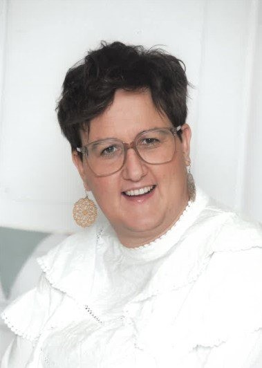 Eine Frau mit kurzen schwarzen Haaren, die eine Brille und ein weißes Hemd trägt, lächelt für die Kamera.
