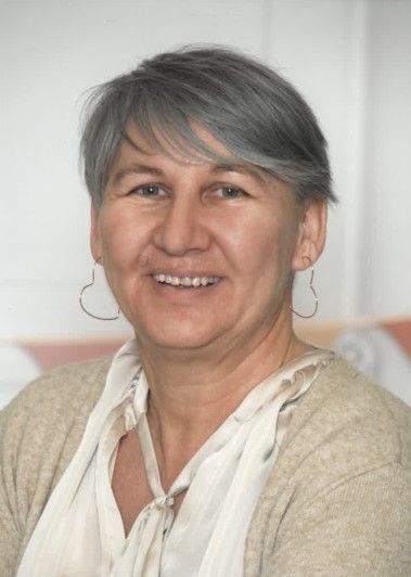 Eine Frau mit grauem Haar lächelt und posiert für ein Foto. Sie trägt ein weißes Blusenshirt und einen beigen Cardigan.