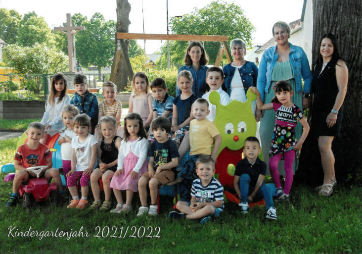 Eine Gruppe von Kindern und Erwachsenen posiert für ein Foto im Garten. Einige sitzen auf dem Rasen, andere stehen hinter ihnen. Eine grüne Puppe ist in der Mitte. Der Text lautet Kindergartenjahr 2021/2022.