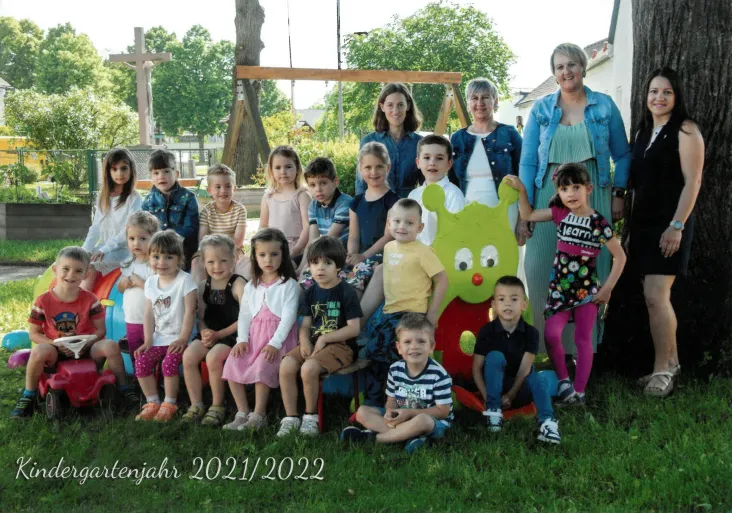 Eine Gruppe von Kindern und Erwachsenen posiert für ein Foto im Garten. Einige sitzen auf dem Rasen, andere stehen hinter ihnen. Eine grüne Puppe ist in der Mitte. Der Text lautet Kindergartenjahr 2021/2022.