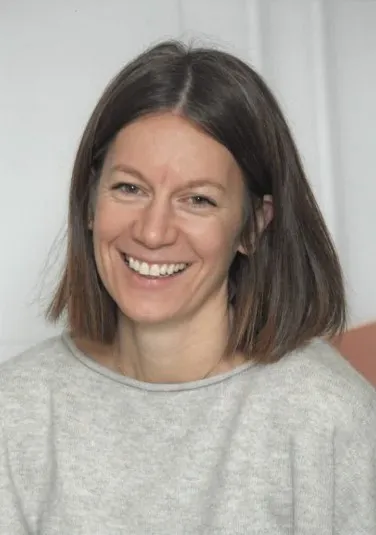 Eine lächelnde Frau mit kurzen braunen Haaren, die ein graues T-Shirt trägt, vor weißem Hintergrund.
