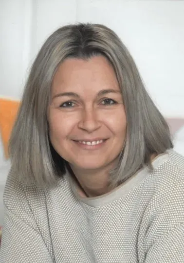 Eine Frau mit grauem Haar lächelt und posiert für ein Foto. Sie trägt ein weißes Oberteil. Hinter ihr ist eine weiße Wand.