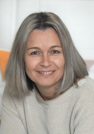 Eine Frau mit grauem Haar lächelt und posiert für ein Foto. Sie trägt ein weißes Oberteil. Hinter ihr ist eine weiße Wand.