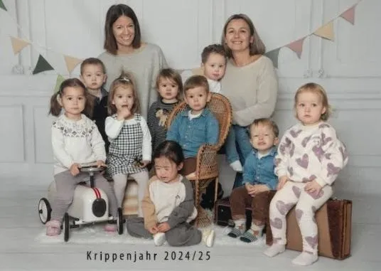Ein Familienfoto mit zwei Frauen, eine sitzt auf einem Stuhl, und mehreren Kindern. Der Text 'Krippenjahr 2024/25' steht unten.