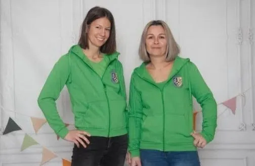 Zwei Frauen stehen Seite an Seite, beide tragen grüne Hoodies mit einem Logo auf der Brust. Sie lächeln und scheinen für ein Foto zu posieren. Der Hintergrund zeigt eine weiße Wand mit Wimpelgirlande.
