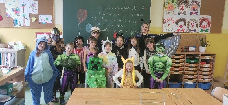 Eine Gruppe von Kindern in verschiedenen Superheldenkostümen steht in einem Klassenzimmer mit einer Lehrerin. Auf der Tafel hinter ihnen steht etwas geschrieben. Es gibt Dekorationen, Regale und einen Tisch im Raum.