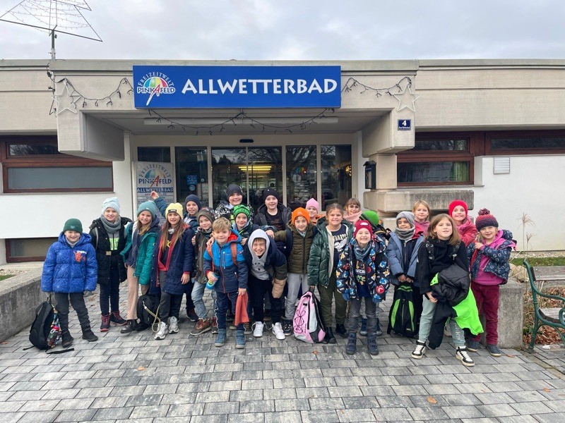 Eine Gruppe von Kindern, alle in Winterkleidung, posiert für ein Foto vor einem Gebäude namens Allwetterbad.