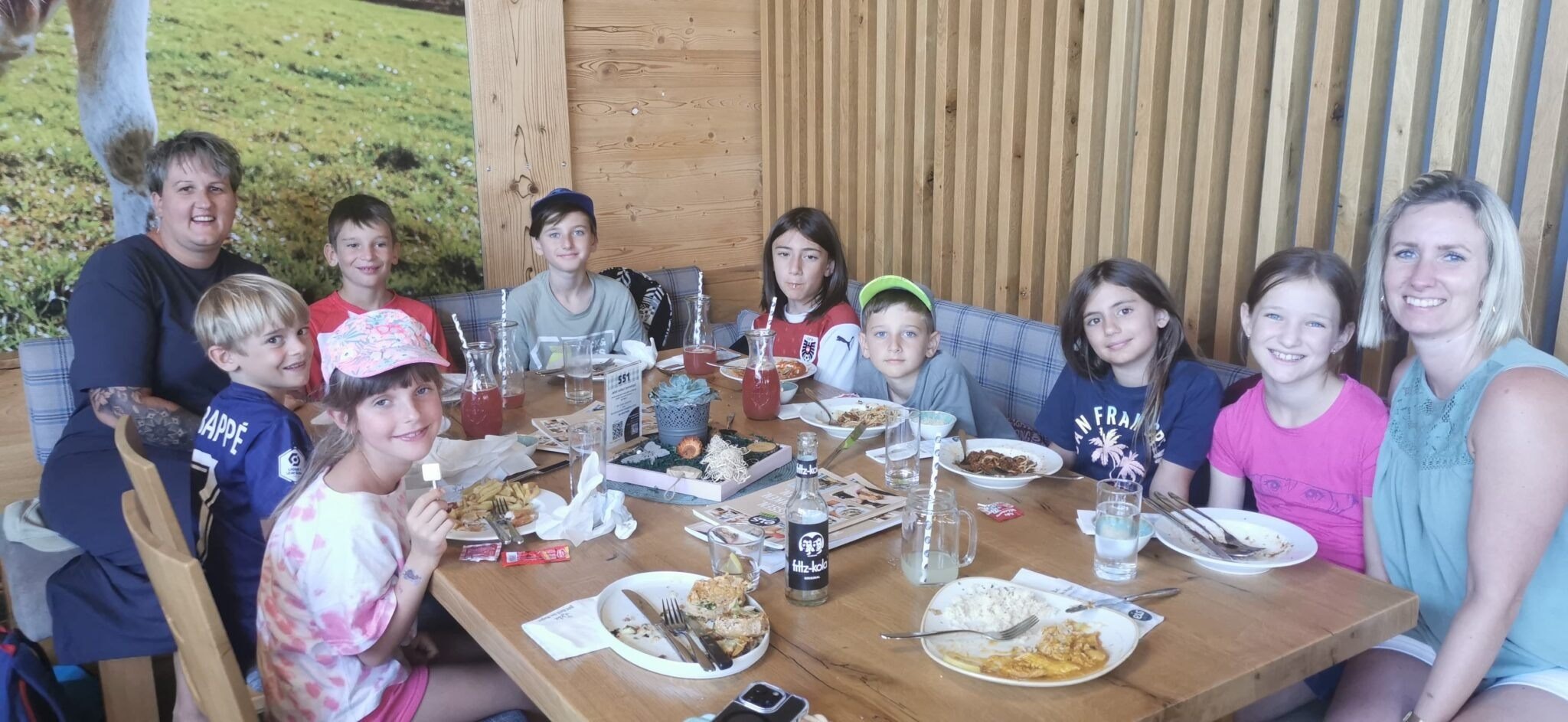 Eine Gruppe von Kindern sitzt um einen Holztisch und posiert lächelnd für ein Foto. Sie haben Teller mit Essen, Getränke und Servietten. Der Tisch ist mit verschiedenen Gerichten, Getränken und Besteck gedeckt. Der Raum hat Holzwände.
