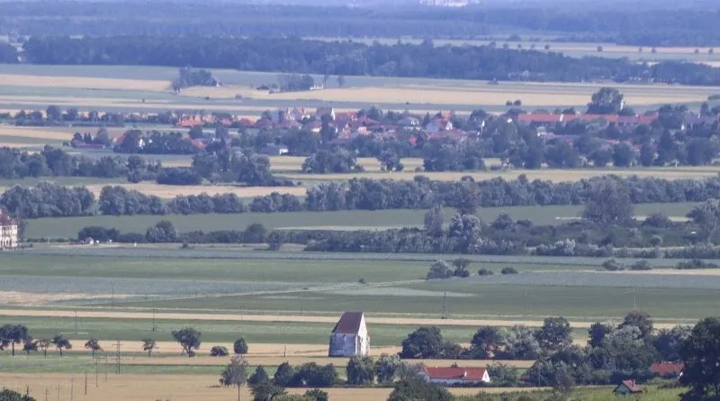 Luftaufnahme einer ländlichen Landschaft mit weiten grünen Feldern, einem kleinen weißen Gebäude und einem Dorf mit roten Dächern in der Ferne.