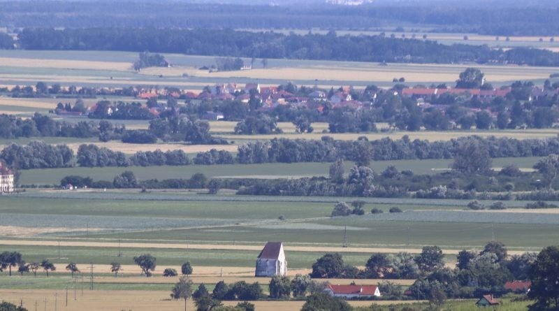 Luftaufnahme einer ländlichen Landschaft mit weiten grünen Feldern, einem kleinen weißen Gebäude und einem Dorf mit roten Dächern in der Ferne.