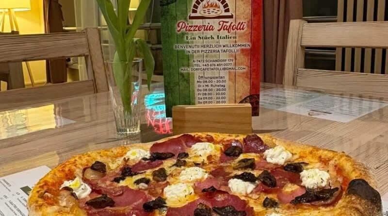 Eine mit verschiedenen Belägen belegte Pizza steht auf einem Holztisch, dahinter eine Blumenvase und ein Menüzettel.